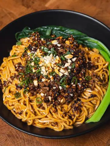 Close-up of Dan Dan Noodles with ground meat, peanuts, and green onions in a black bowl - GENIUS EATS Close-up shot of Dan Dan Noodles with ground meat, peanuts, and green onions in a black bow
