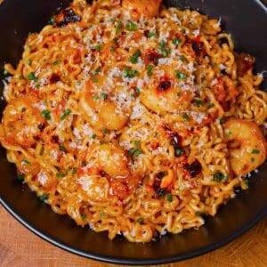 Savory shrimp ramen stir-fry with vegetables, topped with cheese and chopped herbs, served in a black bowl.