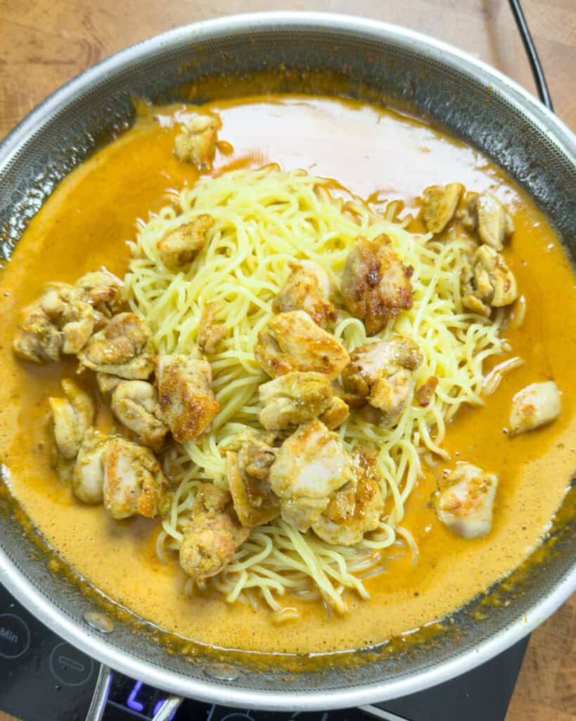 Noodles in orange curry sauce with bite-sized chicken pieces in a shallow pan.